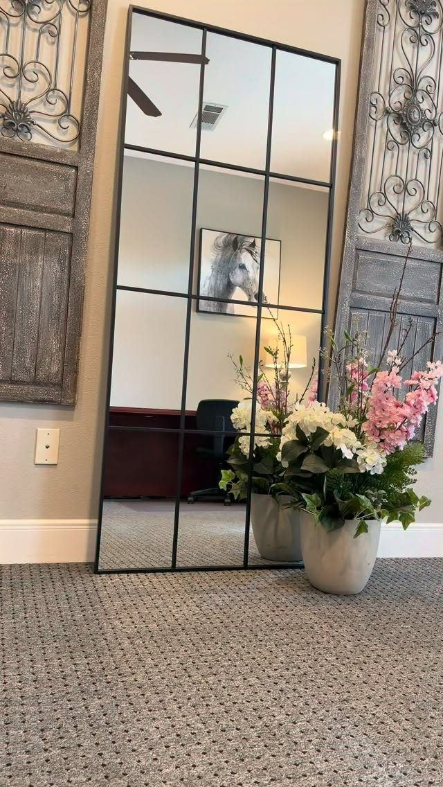 Black windowpane mirror leaning against a wall reflecting a home office and flowers.