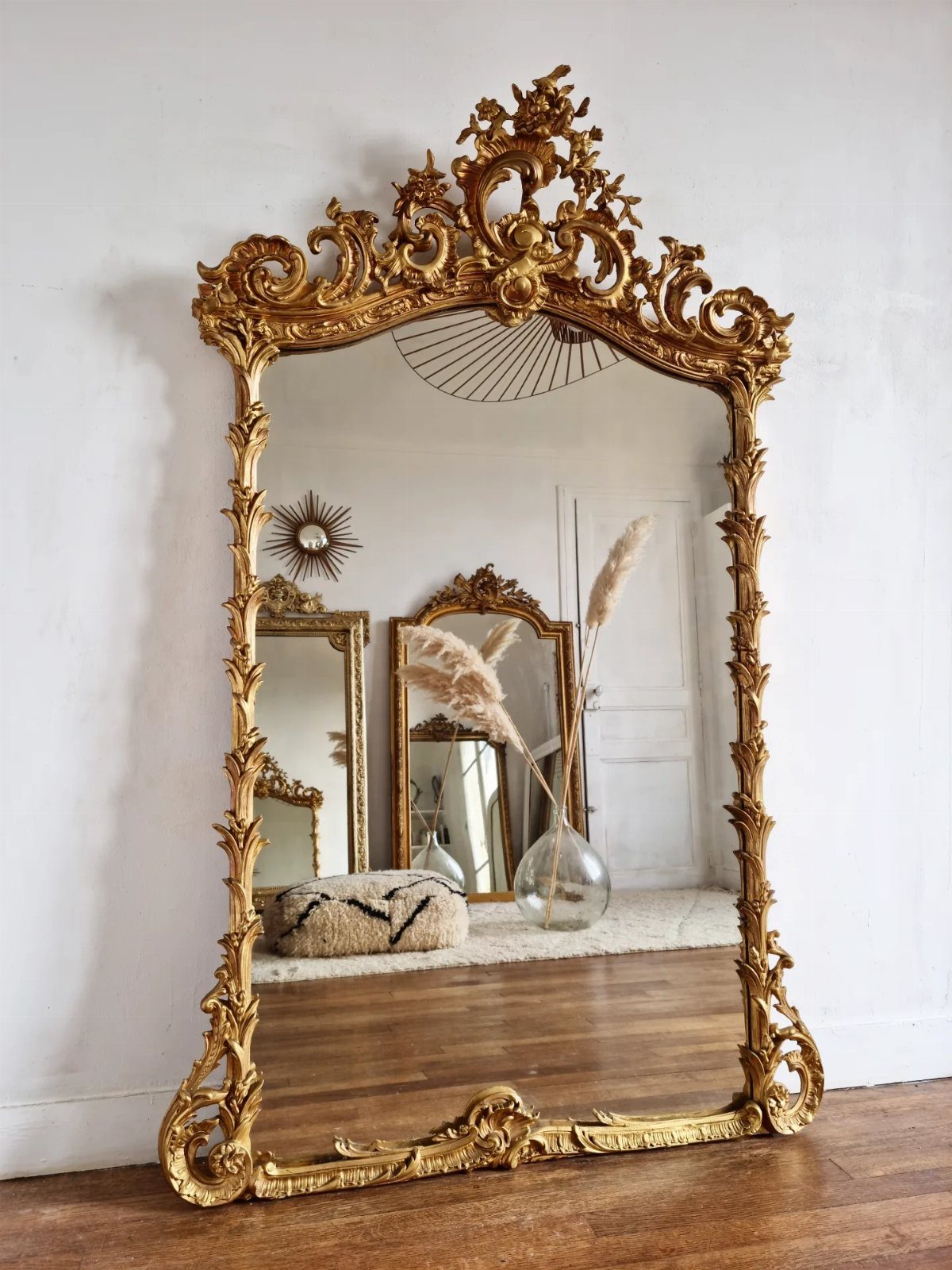 Ornate gilded gold mirror leaning against a white wall on wooden flooring.