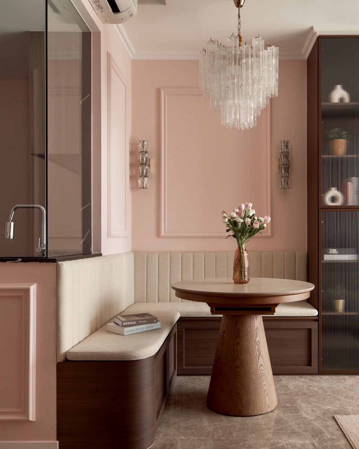 Dining nook with blush pink walls, curved cream banquette, and crystal chandelier.