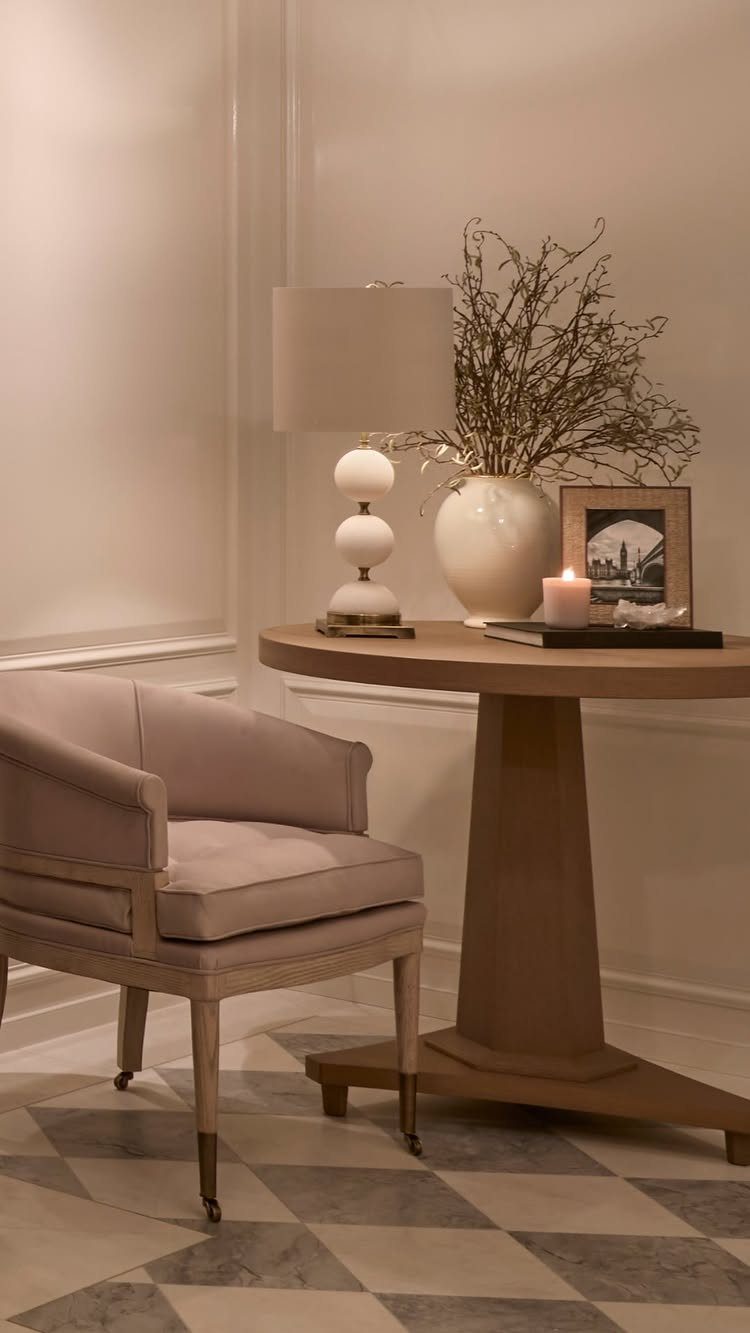 White table lamp on a round wooden table next to a beige armchair.