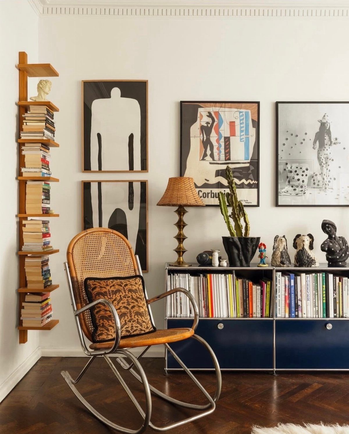Blue sideboard with books and sculptures beneath framed art and a wicker chair.