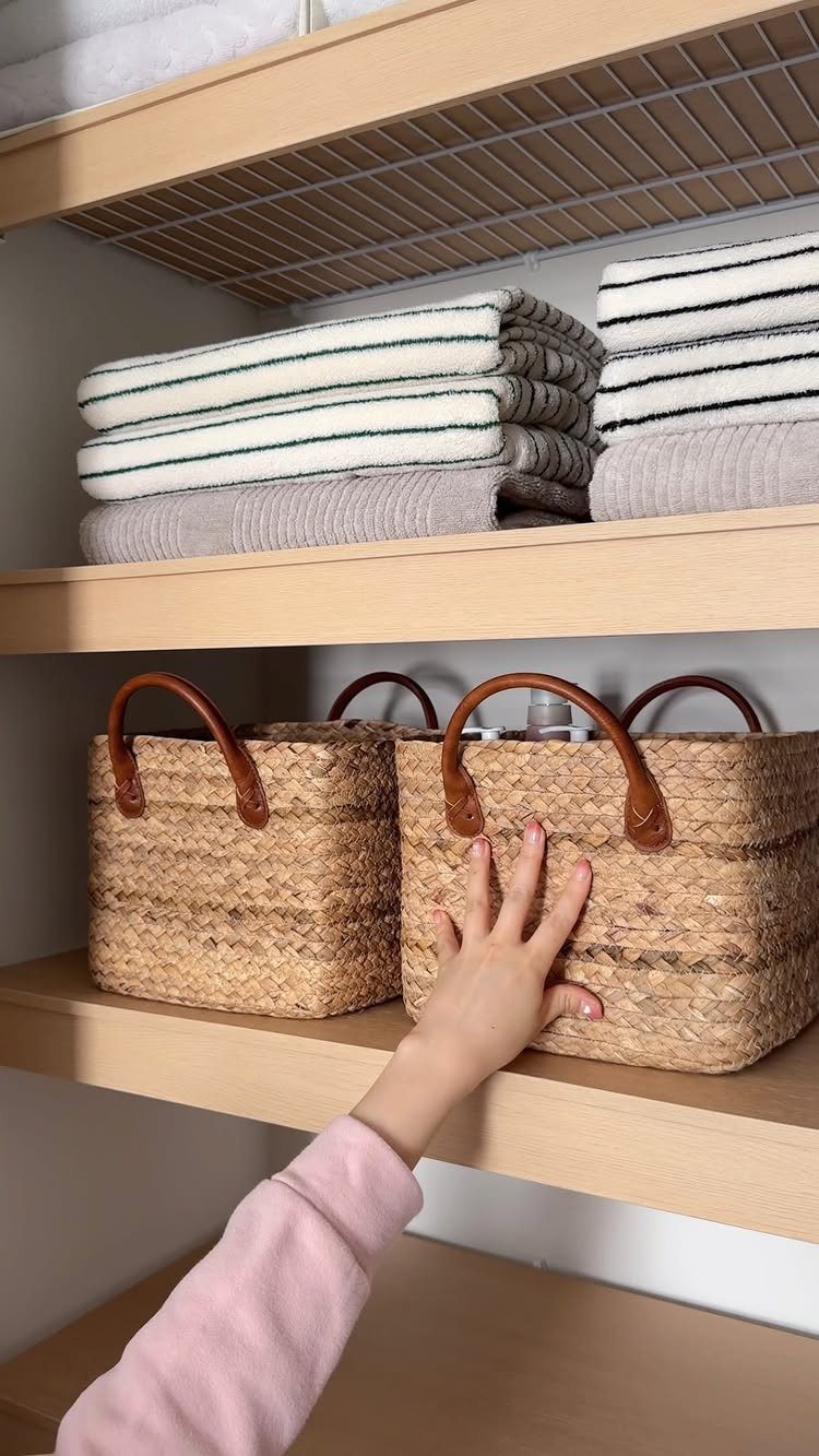 Woven wicker baskets with leather handles on light wood shelves with folded towels.