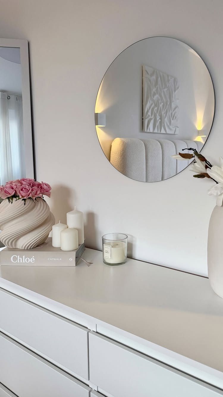 White dresser with a round mirror, pink roses, and white candles in a room.