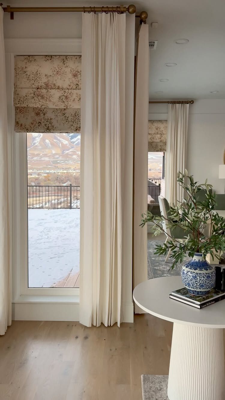 Floral roman shade and cream curtains framing a window with a mountain view.