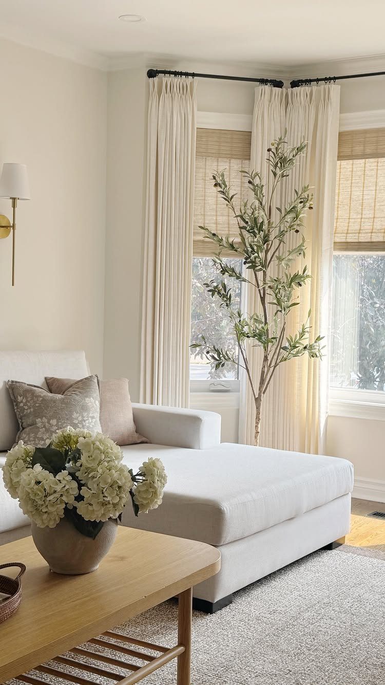White chaise lounge and wooden table in a room with neutral roman shades and curtains.