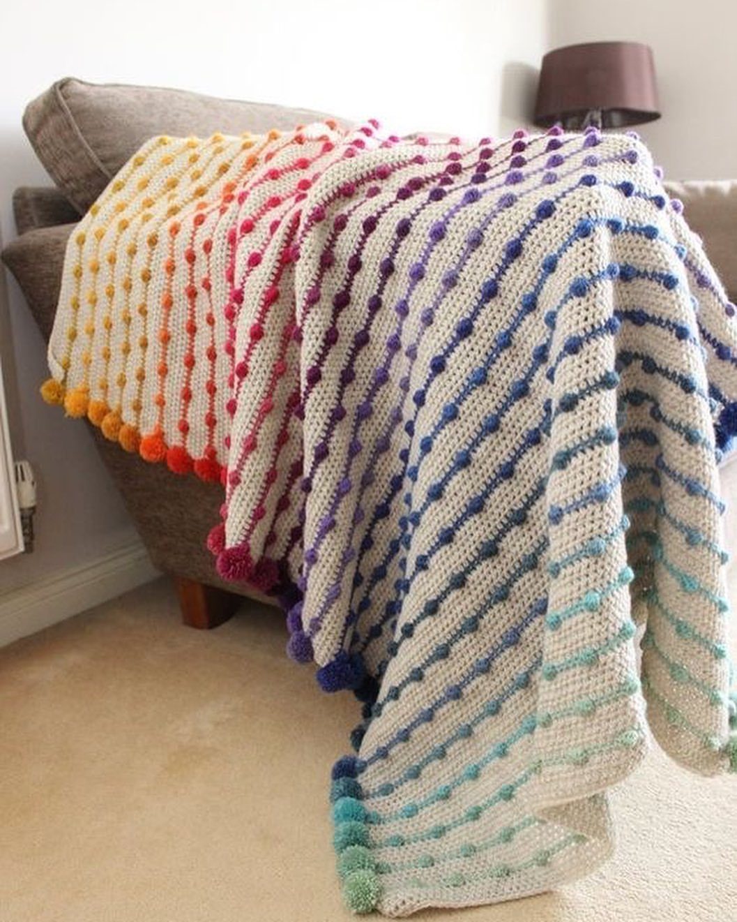 Rainbow crocheted blanket with pom-poms draped over a brown sofa in a room.