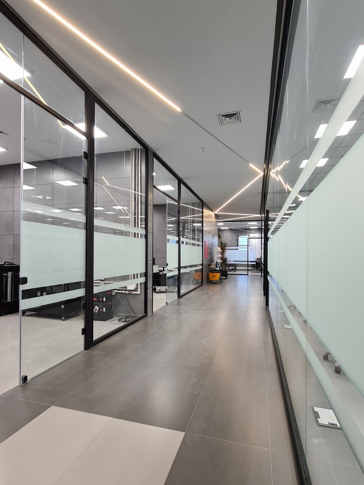 Modern office hallway with glass partition walls and linear ceiling lighting.