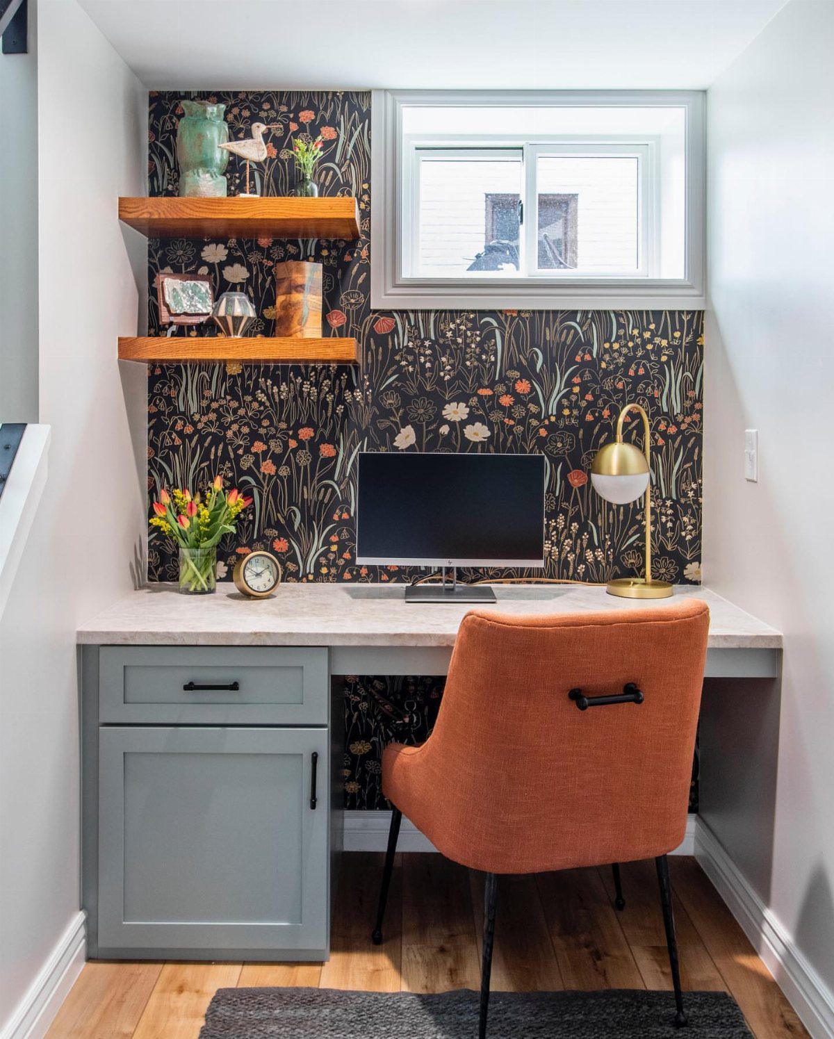 Small office nook with orange chair, grey desk, and dark floral wallpaper.
