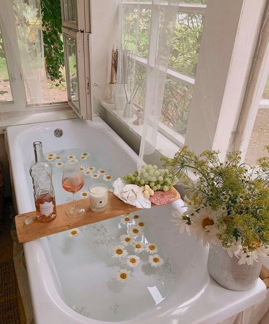 White bathtub with floating daisies, wine on a wooden tray, and fresh flowers.