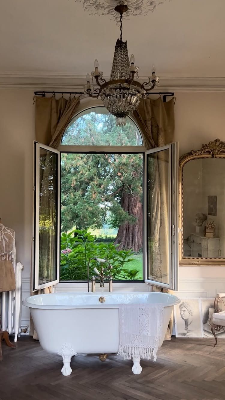 White clawfoot bathtub with gold hardware positioned before an open window overlooking a garden.