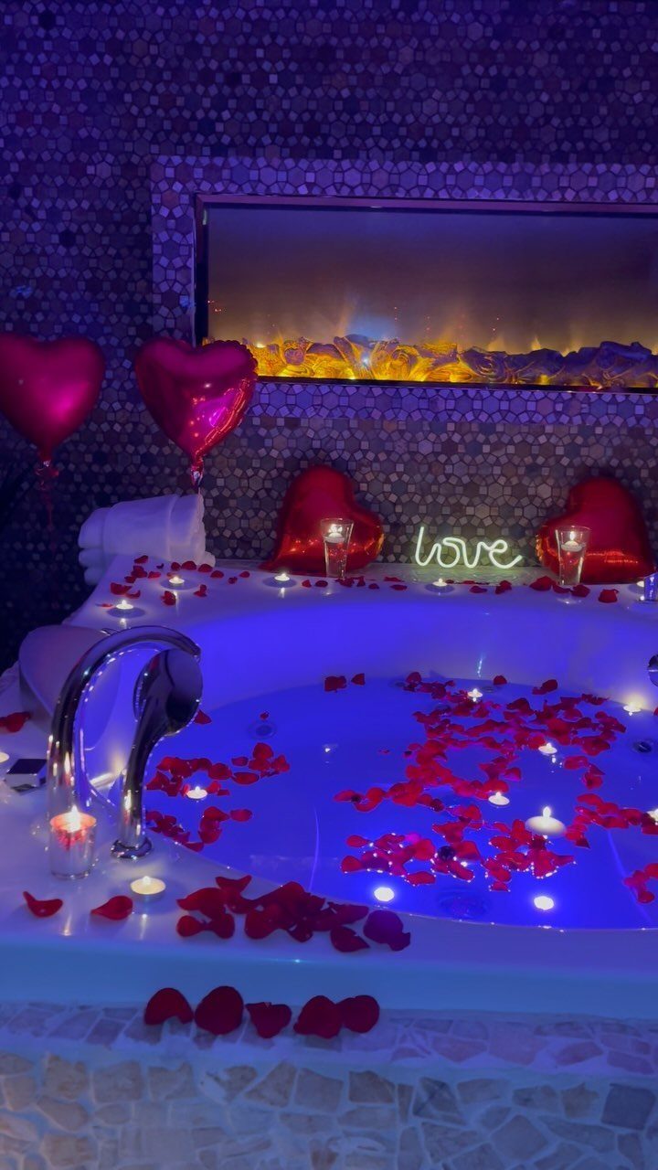 White whirlpool tub with red rose petals, heart balloons, and romantic candle lighting.