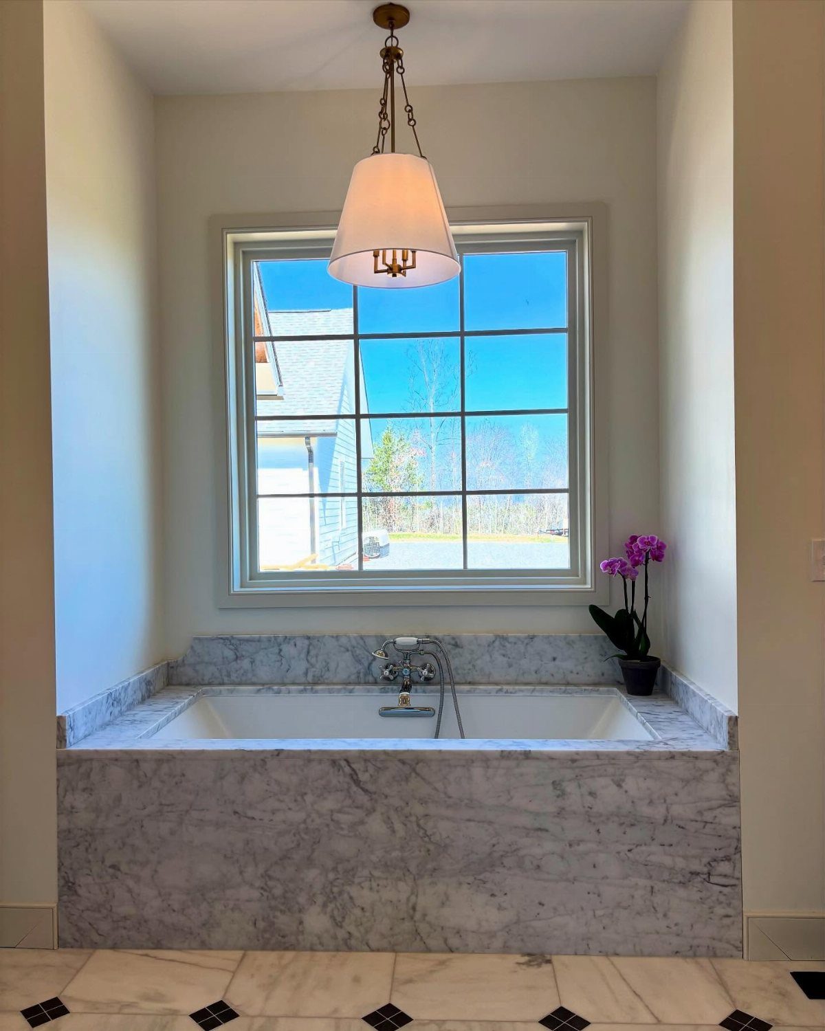 Marble bathtub surround under a grid window with a hanging pendant light and orchid.
