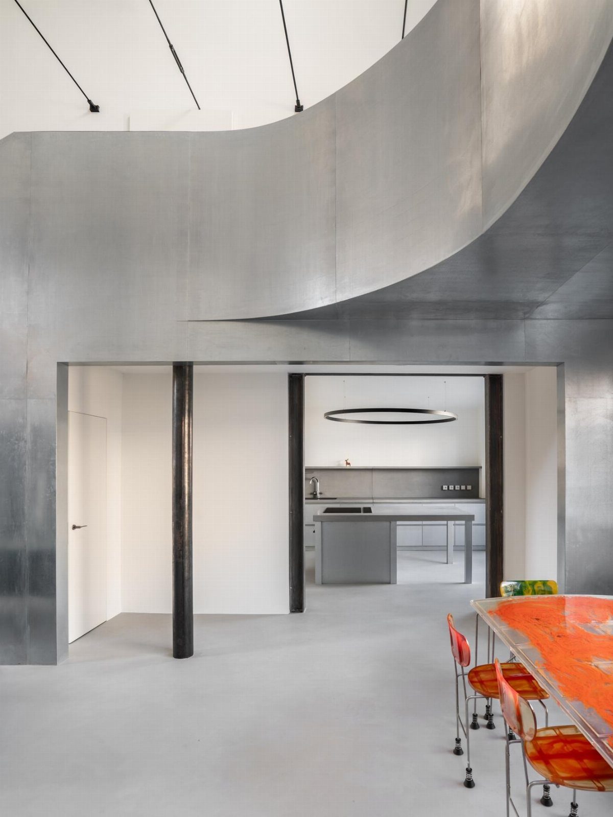 Modern interior with curved metallic silver walls, grey floors, and an orange dining table.