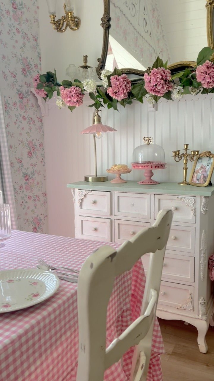 Pink and white vintage interior with checkered tablecloth, ornate dresser, and floral accents.