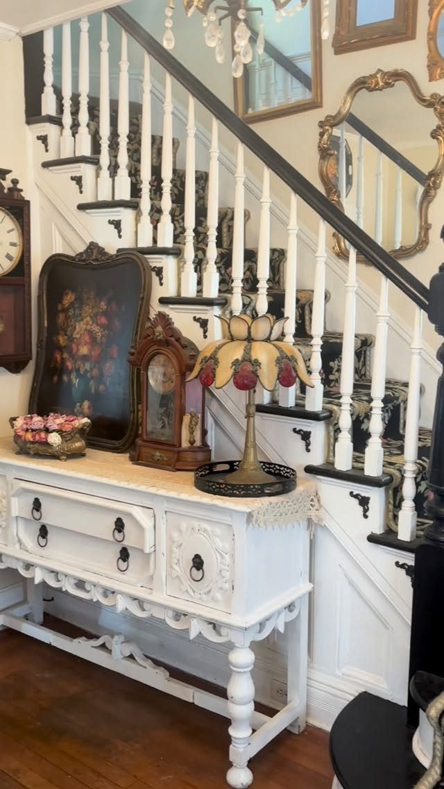 White ornate console table with vintage clock, lamp, and painting under a staircase.