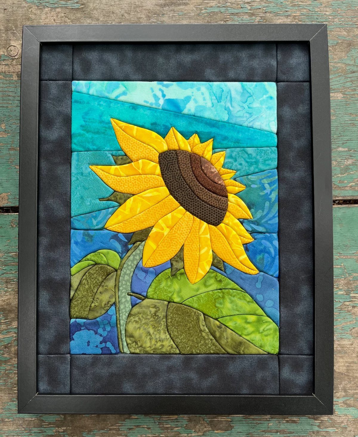 Framed fabric art of a yellow sunflower against a blue textile background.