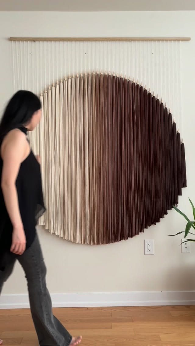 Circular woven wall hanging with brown gradient threads on a white wall.